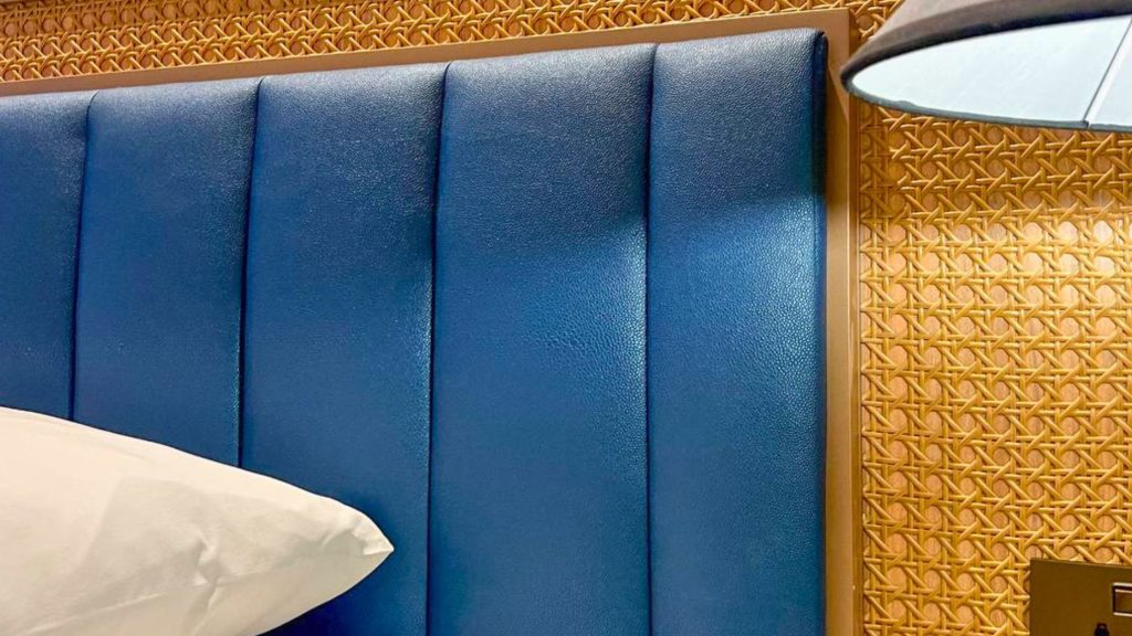 A close-up of a blue upholstered headboard with a woven cane accent wall and a white pillow.
