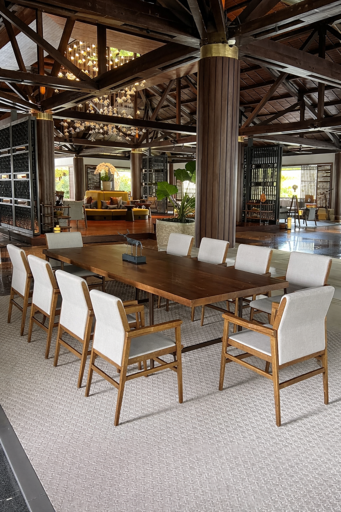 Spacious luxury lounge with a long wooden dining table, upholstered chairs, and exposed timber beams under warm chandelier lighting.