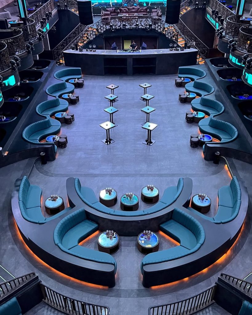 Modern nightclub interior with curved blue seating, glowing tables, and a central DJ booth beneath dramatic lighting.