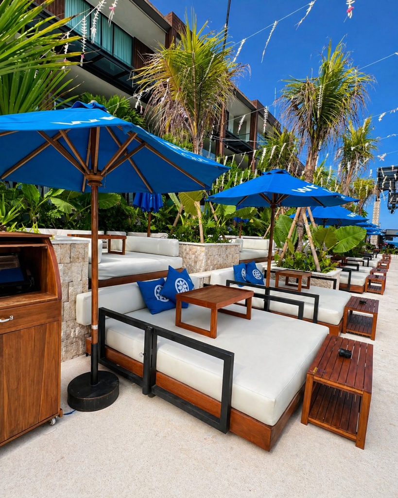 Stylish beachfront lounge seating at White Rock Beach Club Bali with blue umbrellas, tropical plants, and modern daybeds under a clear sky.