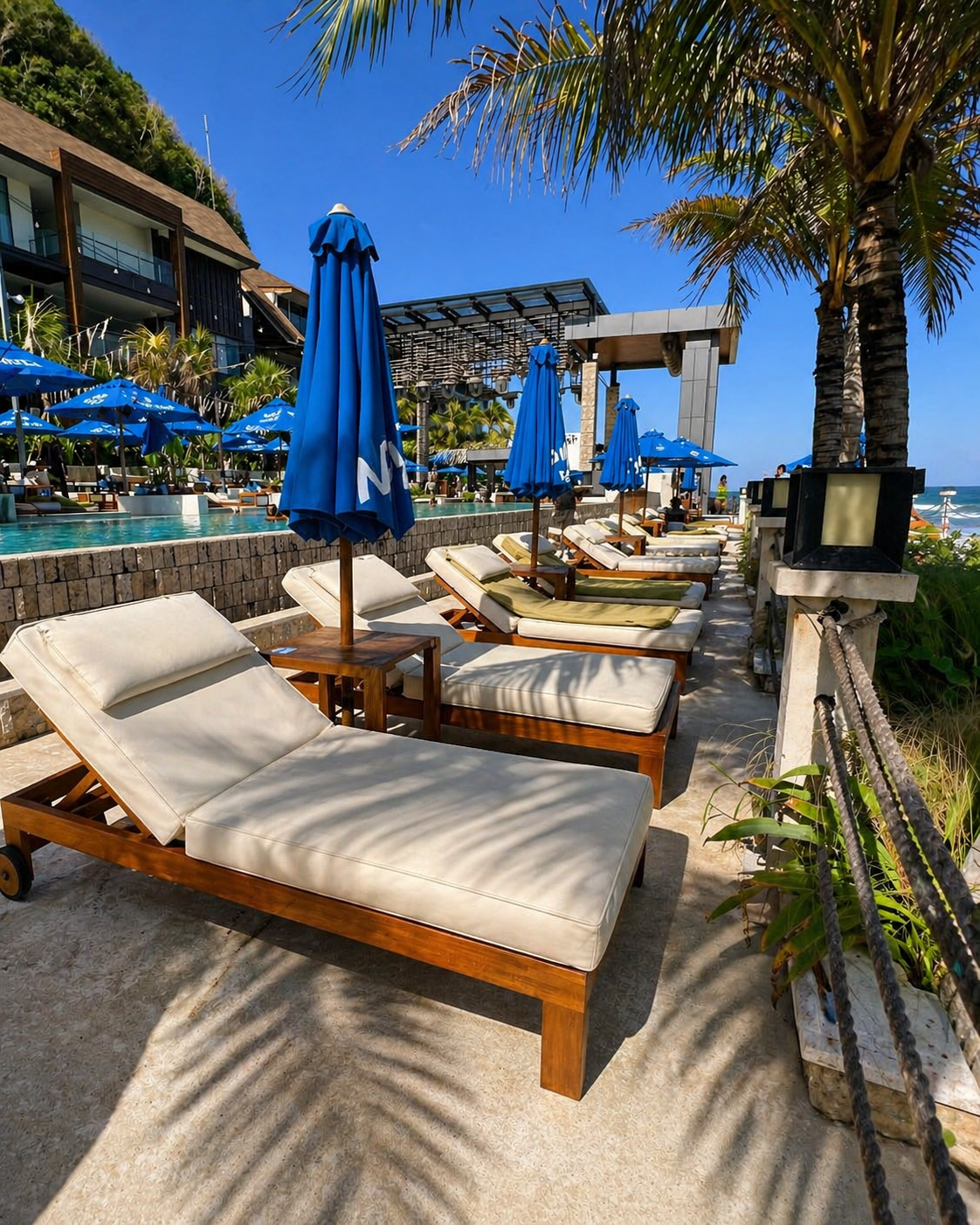 Row of cushioned lounge chairs and blue umbrellas beside a tropical beachfront resort pool under palm trees and a clear blue sky.